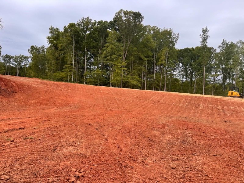 Wide expanse of freshly graded and compacted red clay with a pine treeline behind