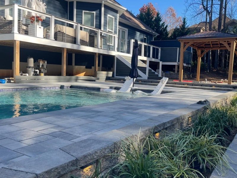 Finished pool with a stone paver patio and pergola beside a lakefront home